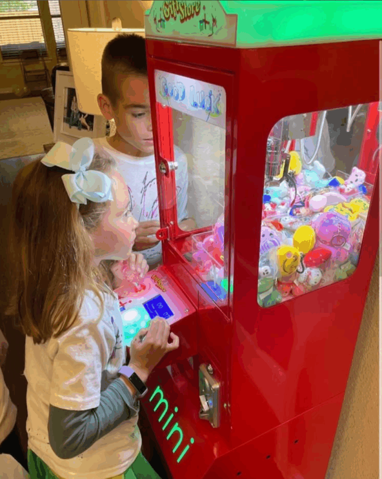 Young girl playing a claw machine at a birthday party with friends watching.